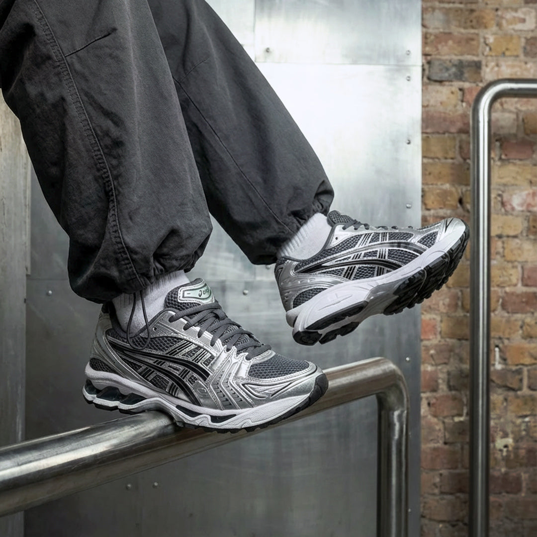 ASICS Gel-Kayano 14 Metropolis Jasper Green on foot resting on metal railing with dark cargo pants showing silver overlays, black tiger stripes, and green tongue branding