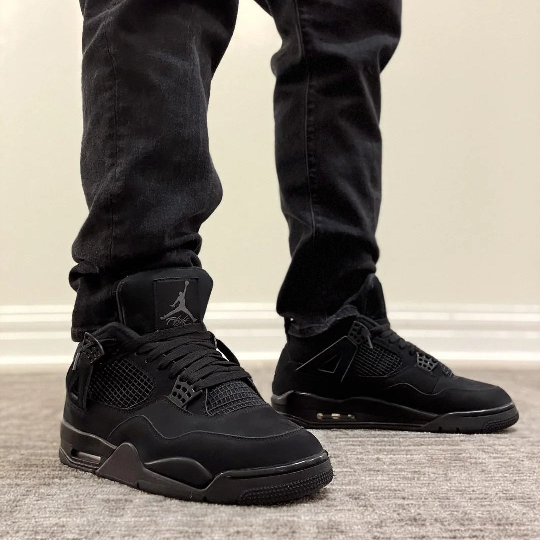 Jordan 4 Retro Black Cat 2025 GS on foot with black jeans front view showing Jumpman Flight tongue logo, all-black nubuck upper, and tonal midsole