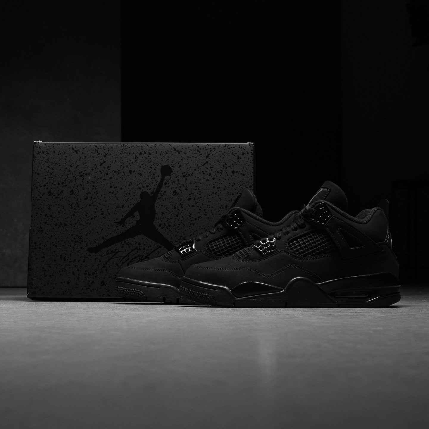 Jordan 4 Retro Black Cat 2025 GS pair displayed with speckled Jordan Brand box in dark studio showing all-black nubuck suede upper and tonal midsole