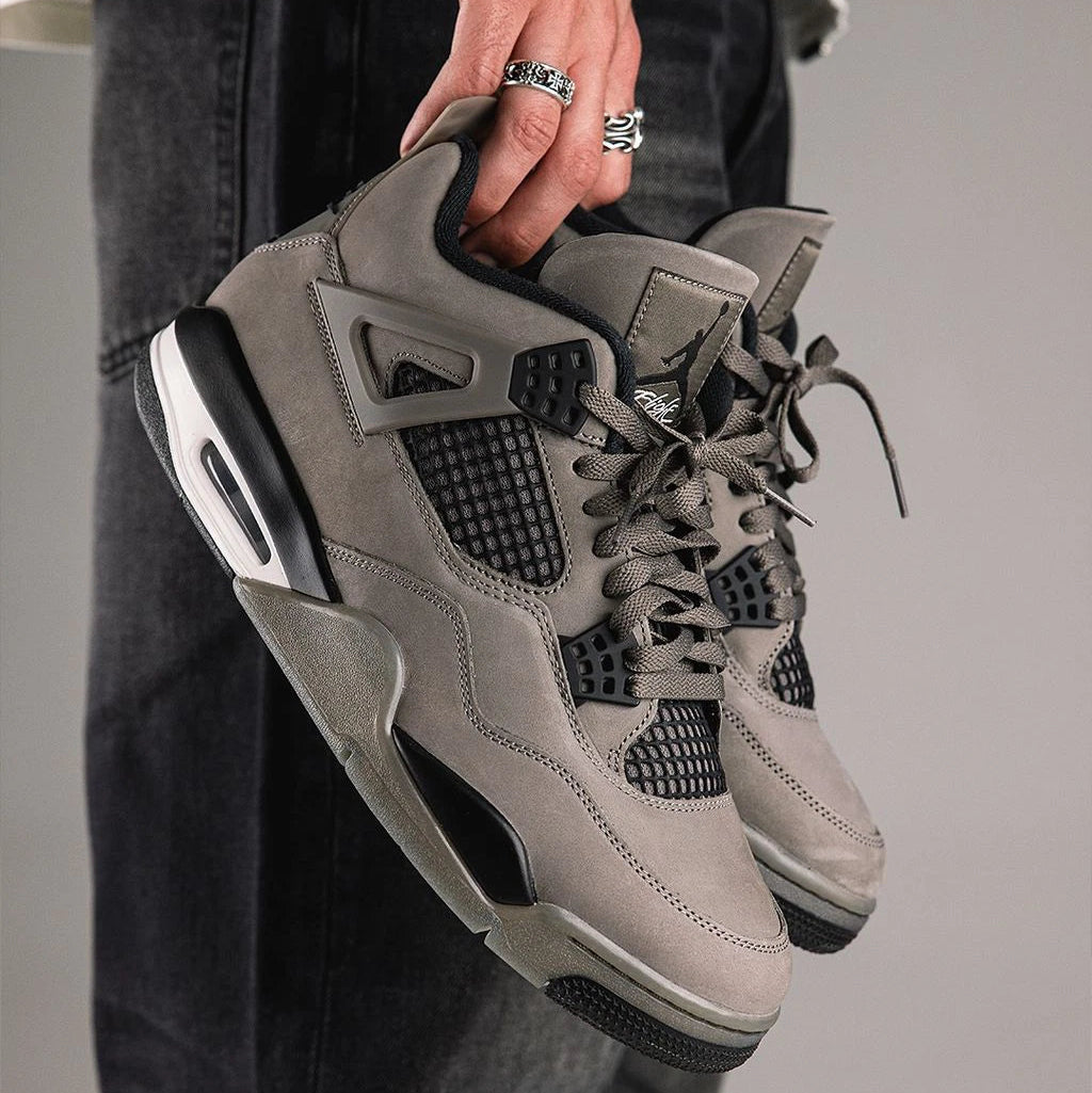 Jordan 4 Retro Cave Stone held in hand by laces with dark denim outfit - showing cave stone brown nubuck upper, black mesh netting, wing eyelets and white Air-Sole unit