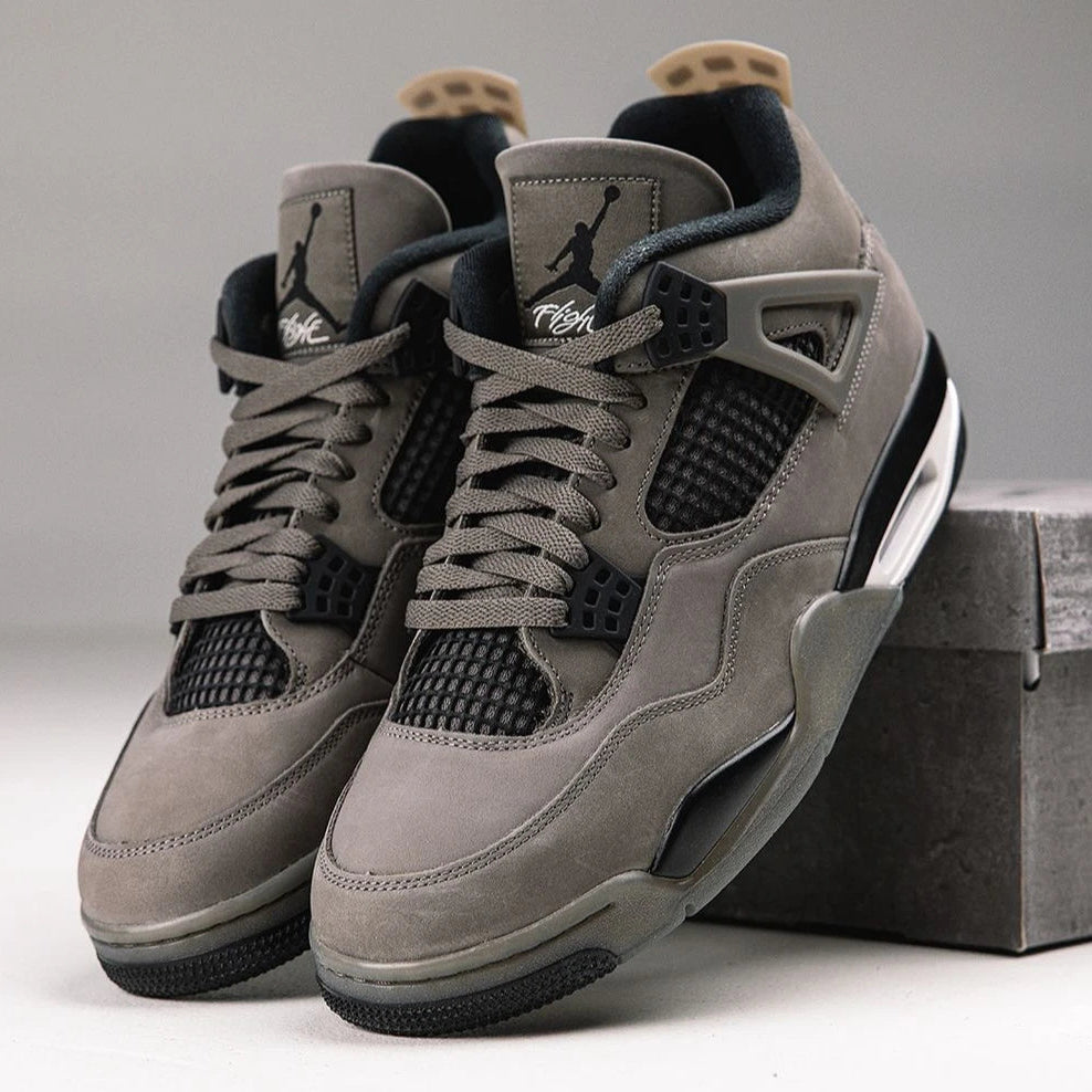 Jordan 4 Retro Cave Stone - front pair view in studio setting with concrete prop showing Jumpman tongue logo, Flight branding, tonal laces, black mesh panels and cave stone nubuck