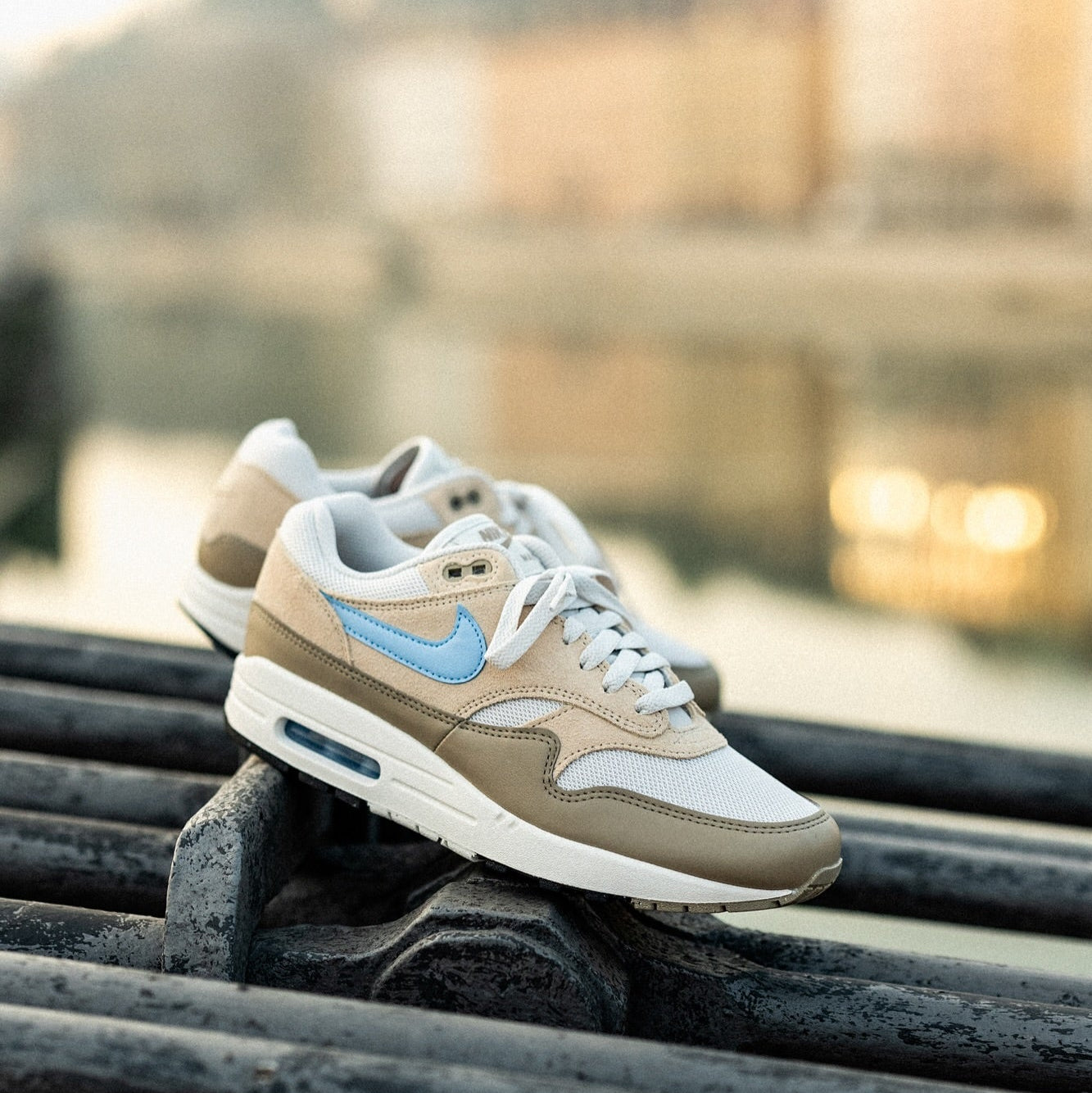 Nike Air Max 1 Light Bone Psychic Blue pair resting on metal railing at golden hour showing light bone suede, blue Swoosh, and white midsole