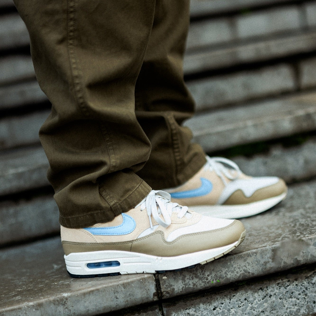 Nike Air Max 1 Light Bone Psychic Blue worn on foot with olive trousers on stone steps showing psychic blue Swoosh, tan suede upper, and visible Air unit