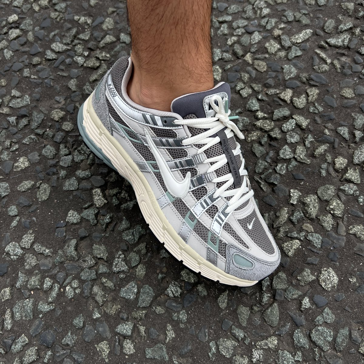 Nike P-6000 Flat Pewter pair held with red Nike shoe box on concrete showing top-down view of grey mesh, silver overlays, and cream midsole