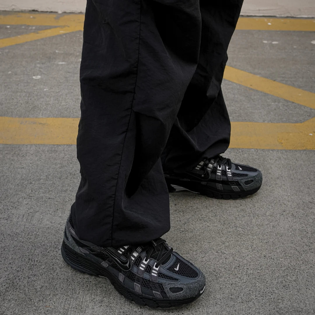 Nike P-6000 Suede Anthracite Metallic Silver worn on foot with black parachute pants on urban pavement showing full shoe silhouette with suede toe and silver accents