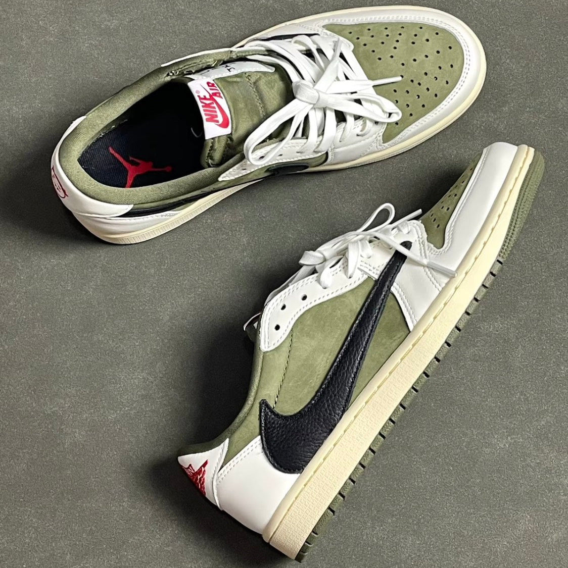 Travis Scott x Air Jordan 1 Low OG Medium Olive - pair held in hands against dark background showing medial view with olive suede, black suede toe, white leather, reverse Swoosh and red Nike Air tongue label
