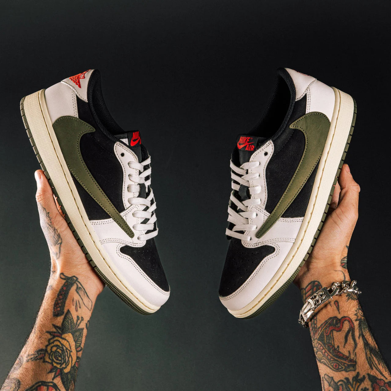 Travis Scott x Air Jordan 1 Low OG Medium Olive - pair held in hands against dark background showing medial view with olive suede, black suede toe, white leather, reverse Swoosh and red Nike Air tongue label