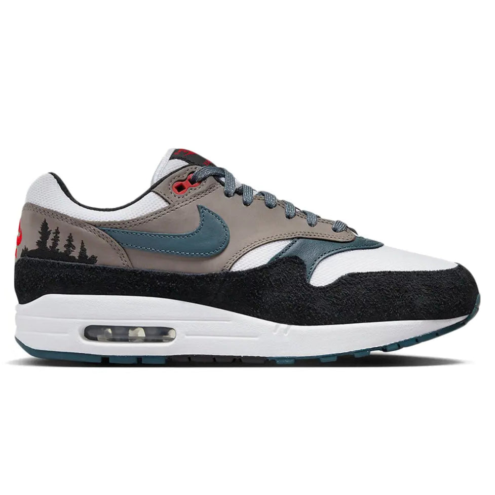 A side view of the Nike Air Max 1 PRM QS Escape trainer, showing the black suede mudguard, Concrete Grey upper, green Nike Swoosh and embroidered treeline detailing.