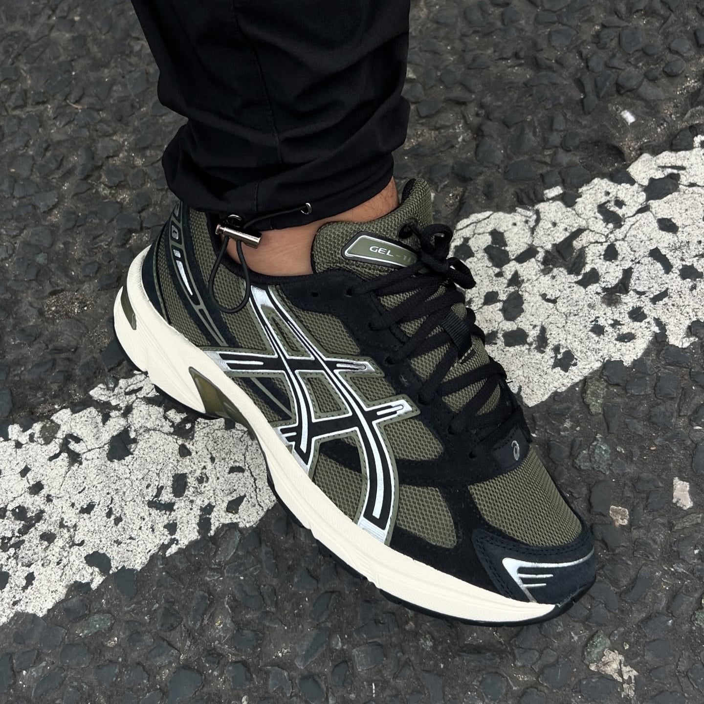 ASICS Gel-1130 Irvine Black on foot top-down view with black trousers on road showing olive green mesh, black overlays, silver stripes, and GEL tongue tag