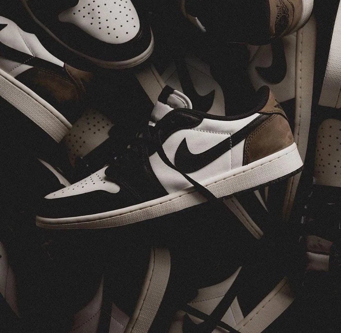 Air Jordan 1 Low OG Mocha multiple pairs stacked in dramatic lighting showing sail leather upper, black Swoosh, and dark mocha nubuck heel panel