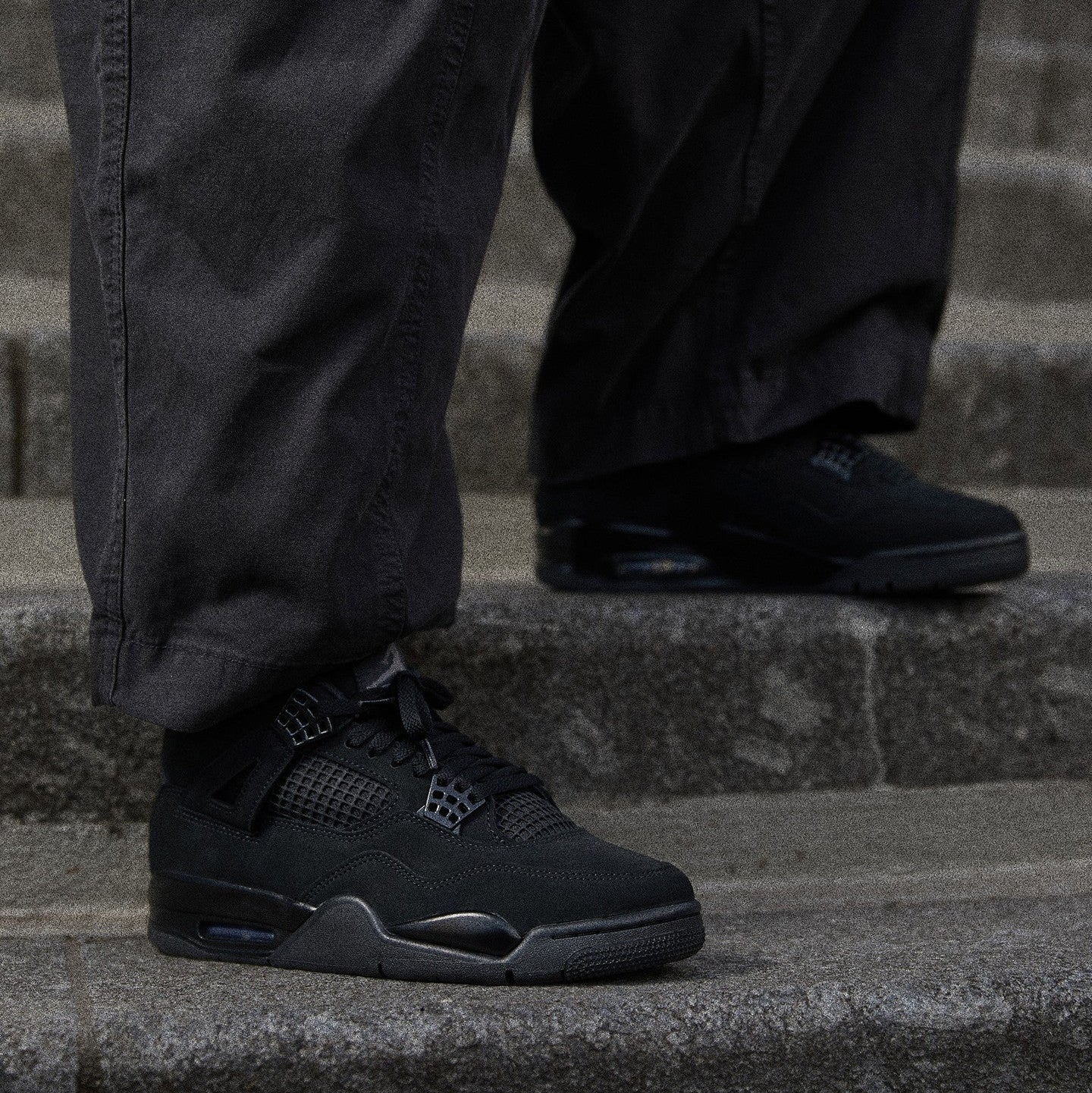Jordan 4 Retro Black Cat 2025 GS worn on foot with dark cargo pants on stone steps showing tonal black nubuck upper, mesh netting, and black TPU cage