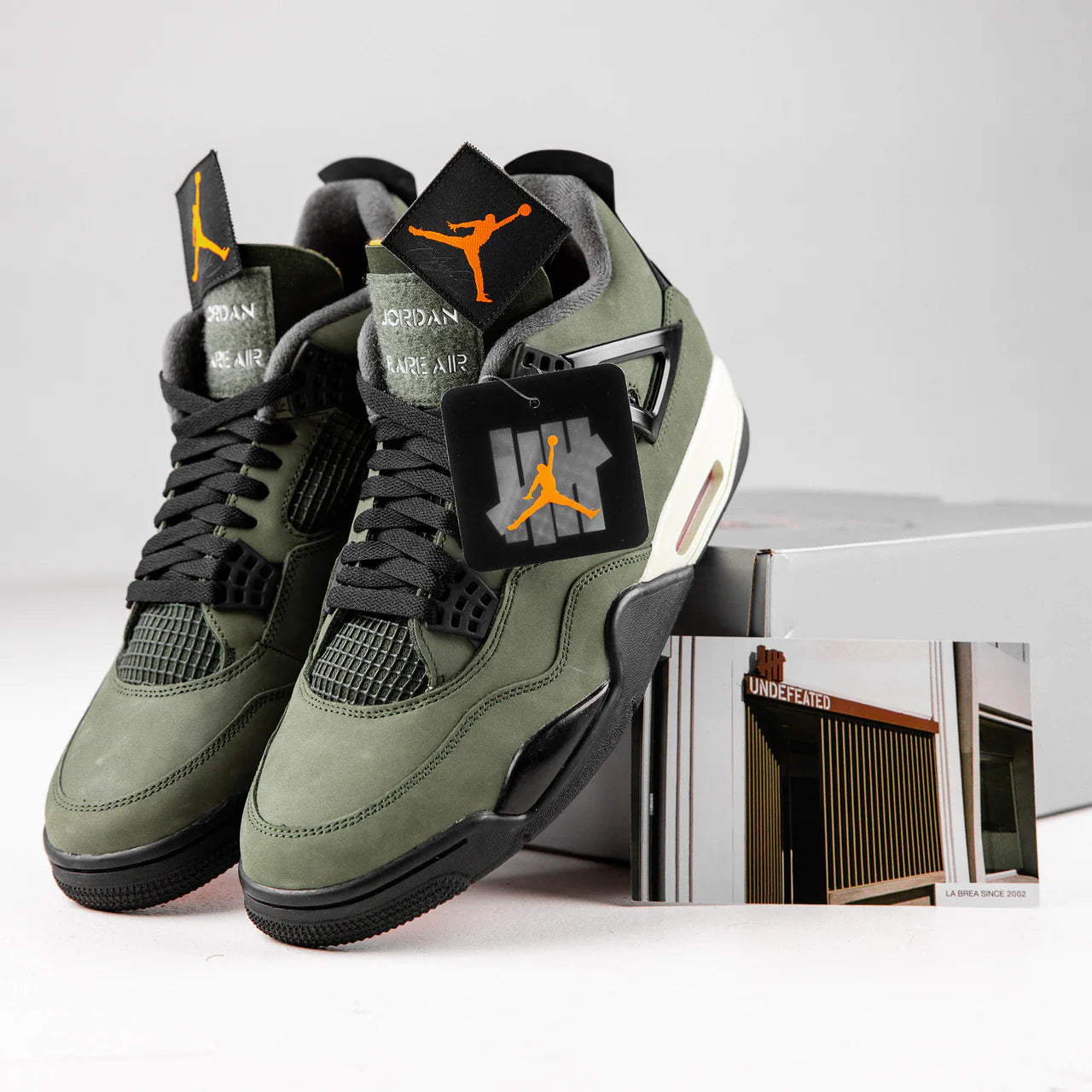 Jordan 4 Retro x UNDEFEATED - pair displayed with orange Jumpman hangtag, UNDEFEATED La Brea postcard and box showing olive suede upper, Jordan Rare Air tongue branding and black laces