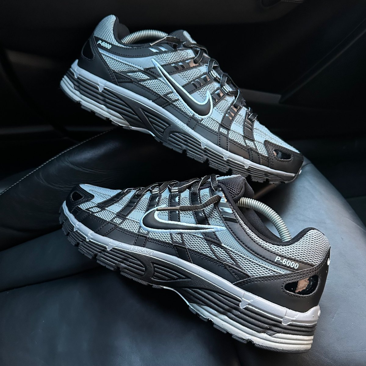 Nike P-6000 Black Cool Grey pair stacked on dark leather seat showing cool grey mesh upper and black synthetic overlays