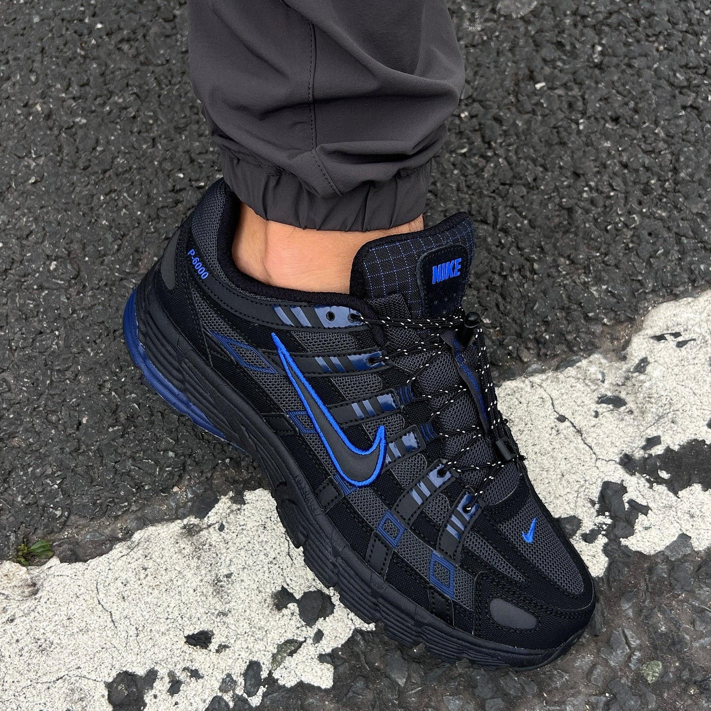Nike P-6000 Black Obsidian on foot on asphalt showing blue Swoosh outline and dark mesh upper with grey trousers