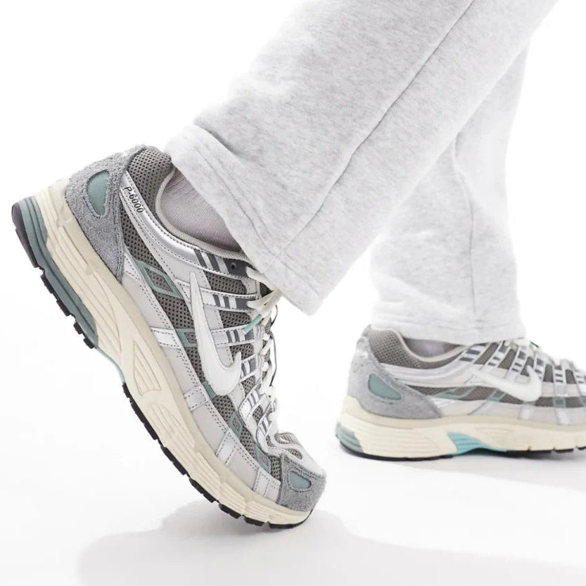 Nike P-6000 Flat Pewter close-up on foot with grey sweatpants showing grey mesh upper, metallic silver straps, white Swoosh, Bowerman Series tongue tag, and aqua midsole accents