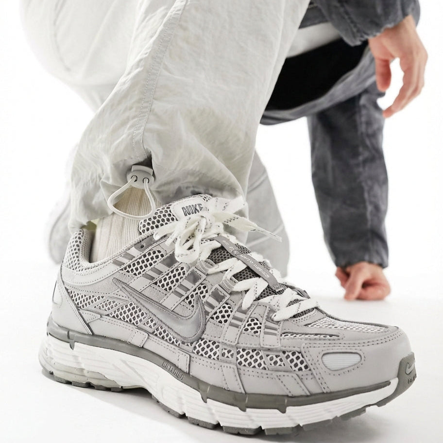 Nike P-6000 Light Iron Ore on foot with white nylon pants showing medial side close-up with grey mesh, leather overlays, silver Swoosh, and perforated toe
