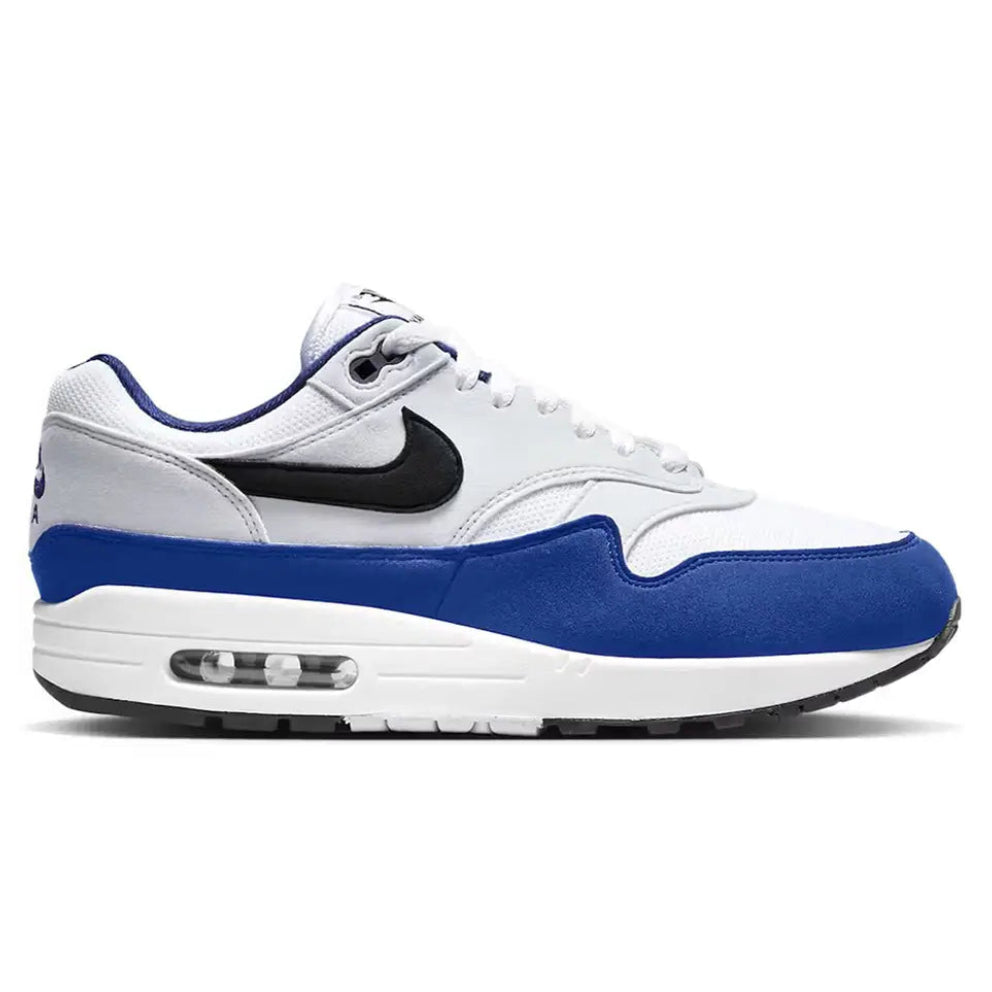 A side view of the Nike Air Max 1 Deep Royal Blue trainer, showing the white mesh upper, blue mudguard, black Swoosh and classic visible Air midsole.