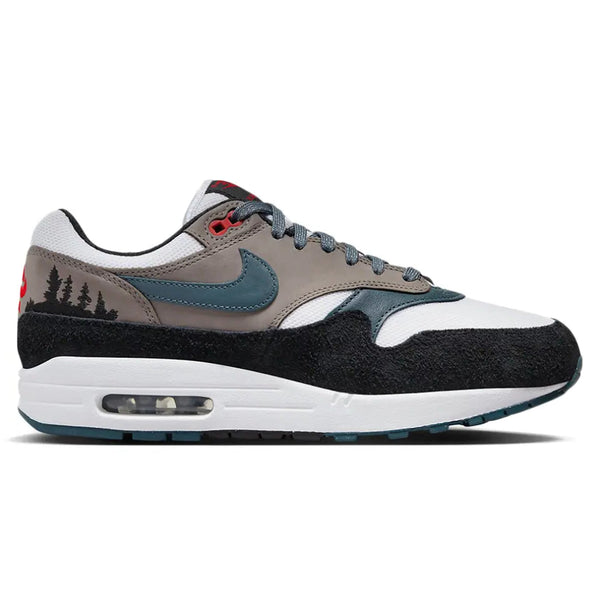 A side view of the Nike Air Max 1 PRM QS Escape trainer, showing the black suede mudguard, Concrete Grey upper, green Nike Swoosh and embroidered treeline detailing.